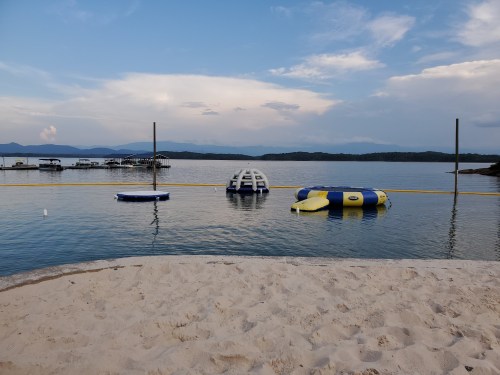A calm lake scene with inflatable water toys, a sandy beach, and distant boats under a blue sky with clouds.