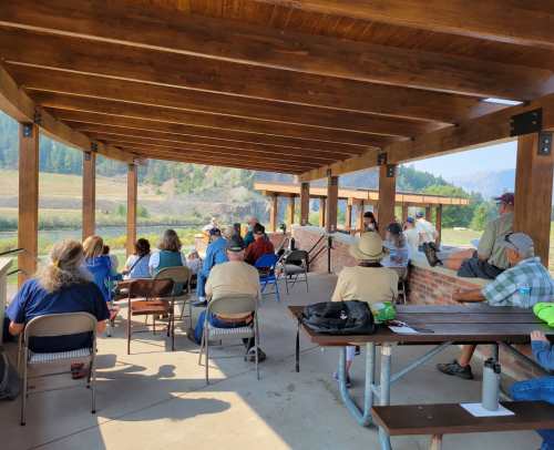 A group of people seated under a wooden pavilion, listening to a speaker with a scenic landscape in the background.