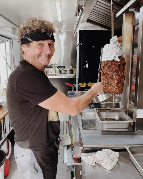 A smiling chef in a food truck holds a large skewer of meat, preparing to serve it.