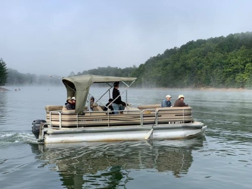 A pontoon boat with several people cruising on a misty lake surrounded by green trees.