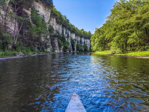 A serene river scene with lush green cliffs and trees, viewed from the front of a canoe.