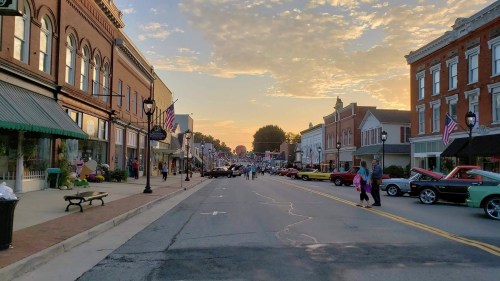 A charming small town street at sunset, lined with shops and classic cars, bustling with people and American flags.