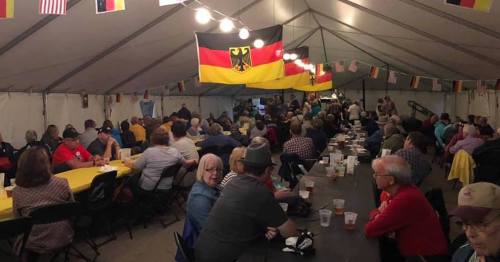 A large gathering under a tent, with people seated at long tables, decorated with German flags and festive lighting.