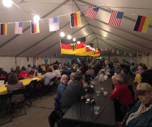 A large gathering under a tent, with people seated at long tables, decorated with flags and string lights.