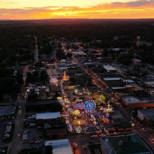 Aerial view of a vibrant carnival at sunset, with colorful lights and rides amidst a small town landscape.