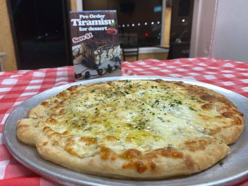 A cheesy pizza on a silver platter with a promotional sign for tiramisu in the background, set on a checkered tablecloth.