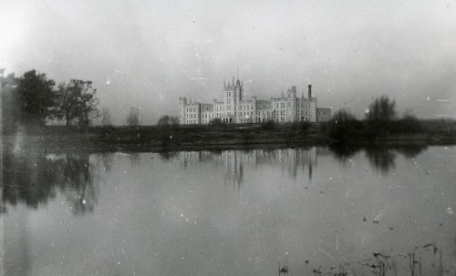 A historic building reflected in a calm lake, surrounded by trees and a misty atmosphere. Black and white photo.