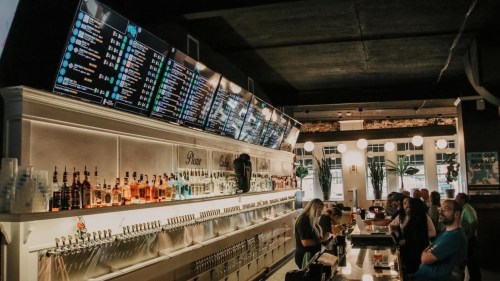 A bar with a long counter, shelves of liquor, and digital screens displaying drink options. Patrons are seated and ordering.