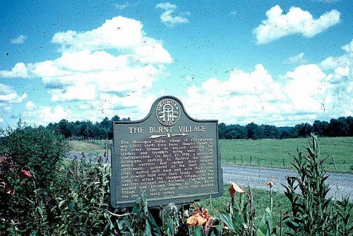 A historical marker titled "The Burnt Village" stands in a grassy field under a blue sky with fluffy clouds.