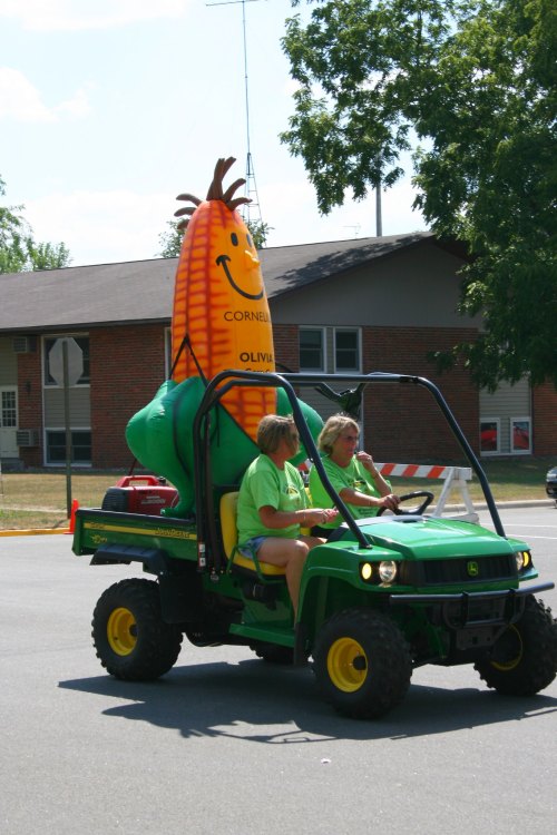 Two women in green shirts ride a John Deere vehicle, featuring a large inflatable corn character in the back.