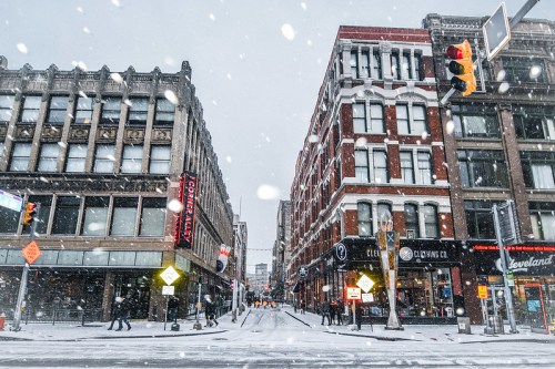 Snow falls on a city street lined with historic buildings, traffic lights, and pedestrians braving the winter weather.