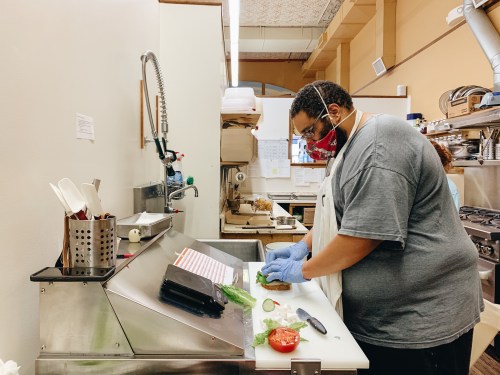 A person in a gray shirt and mask prepares food at a kitchen counter, wearing gloves and chopping vegetables.