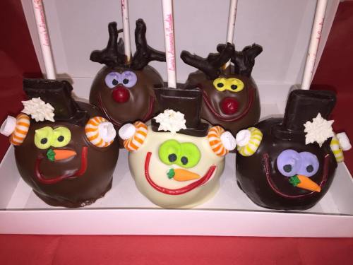 Five decorated chocolate-covered apples resembling festive characters: reindeer, snowman, and a turkey, with colorful candy accents.