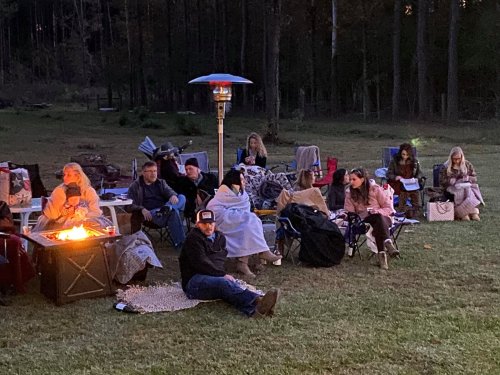 A group of people sitting around a fire pit in a grassy area, wrapped in blankets, enjoying an outdoor gathering.