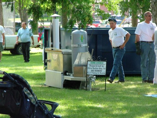 A corn cooker setup at an outdoor event, with people in the background and a sign detailing its purpose.