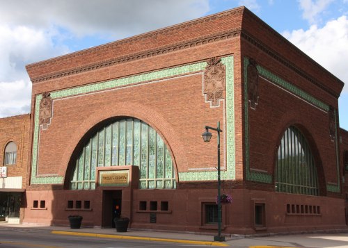Historic brick building with large arched windows and decorative green tiles, set against a blue sky.