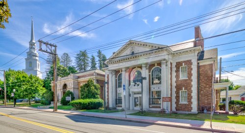 Historic building with columns, surrounded by greenery, and a church steeple visible in the background under a clear blue sky.