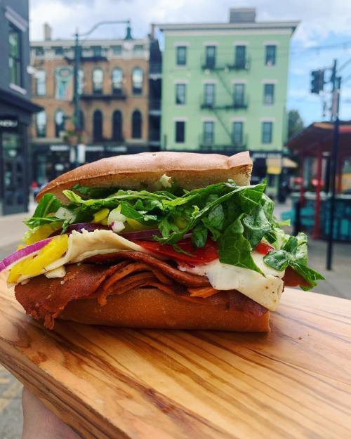 A close-up of a sandwich loaded with lettuce, tomatoes, onions, and deli meat, held on a wooden platter outdoors.