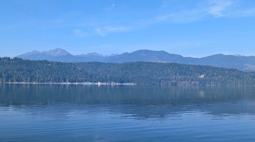 A serene lake reflecting mountains and a clear blue sky, surrounded by lush green trees.