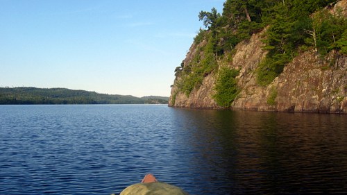 A serene lake scene with calm waters, rocky cliffs, and lush greenery under a clear blue sky.