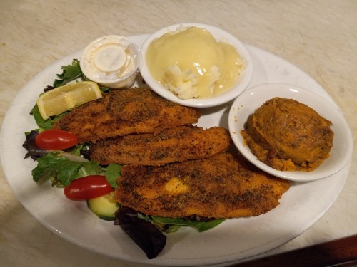 A plate with three fried fish fillets, mashed potatoes, a side of coleslaw, and fresh vegetables.