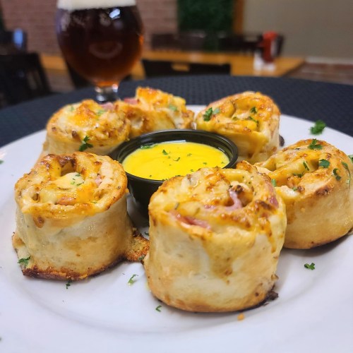 A plate of cheesy, rolled-up snacks with a side of dipping sauce, garnished with parsley and a beer in the background.