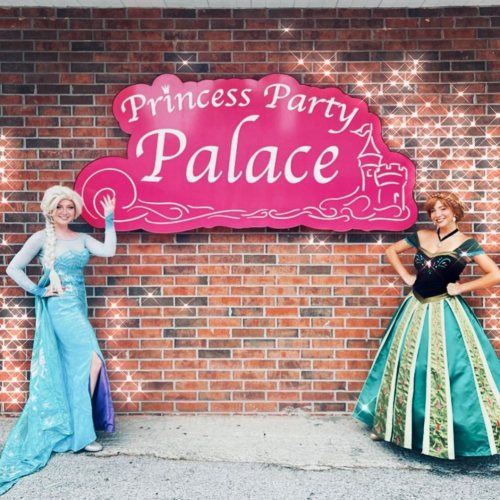 Two costumed characters resembling princesses stand beside a pink sign that reads "Princess Party Palace" against a brick wall.