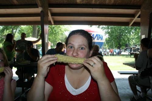 A woman in a red shirt smiles while holding a corn on the cob, sitting at a picnic table in a park.