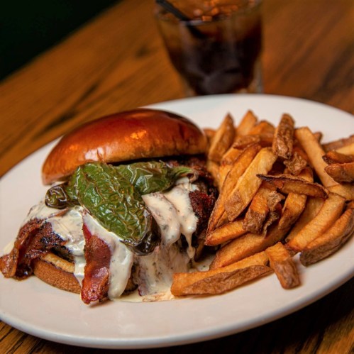 A juicy burger topped with bacon, jalapeños, and creamy sauce, served with crispy fries and a drink in the background.
