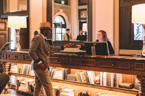 A man in a hat interacts with a smiling receptionist at a stylish hotel lobby. Bookshelves are visible in the background.