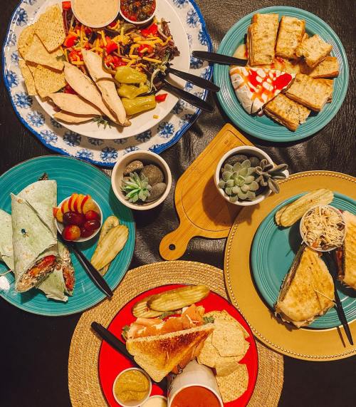 A colorful spread of various dishes, including wraps, sandwiches, chips, and dips, arranged on vibrant plates.