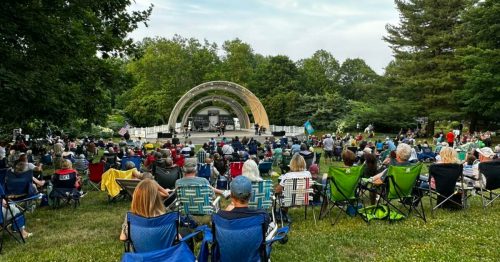 A large crowd sits on lawn chairs in a park, watching a performance on a stage surrounded by trees.