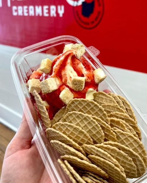 A hand holding a container of strawberry ice cream topped with marshmallows, alongside a stack of waffle cookies.