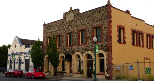 Historic stone building with arched windows, flanked by a white building, and parked red cars in front.