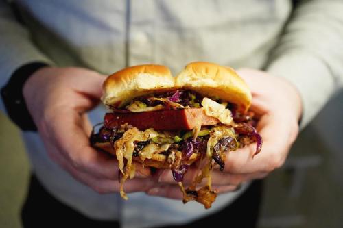 A person holds a sandwich filled with colorful slaw, grilled vegetables, and a thick slice of meat between two soft buns.
