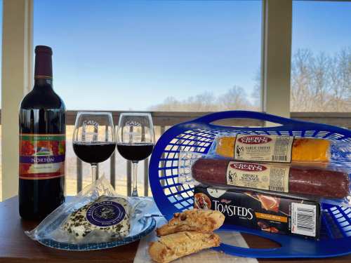 A bottle of red wine, two glasses, cheese, salami, crackers, and biscotti on a table with a scenic view.