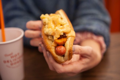 A person holds a hot dog topped with macaroni and cheese, with a drink in the background.