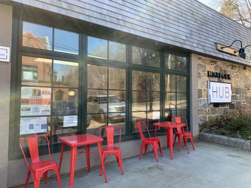 Exterior of Norfolk Hub with large windows, a stone wall, and red tables and chairs outside.