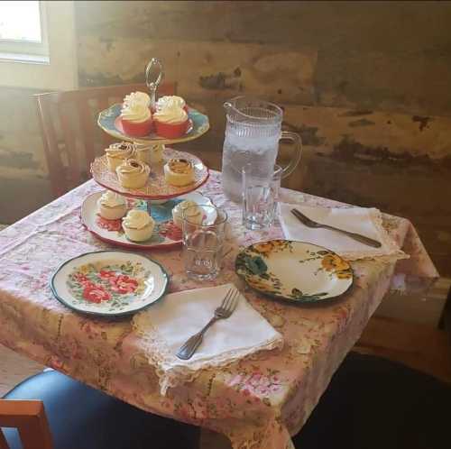 A cozy table set with floral plates, glasses, and a tiered stand of colorful cupcakes, against a rustic wall.