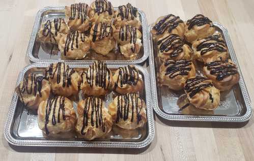 Three trays of cream puffs drizzled with chocolate sauce, arranged neatly on a wooden surface.