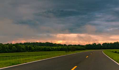 A winding road through lush green fields under a dramatic sky with clouds and a hint of sunset.