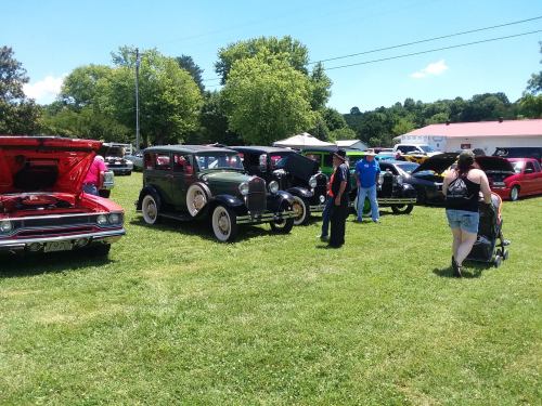 A car show featuring classic cars on a sunny day, with attendees admiring the vehicles in a grassy area.
