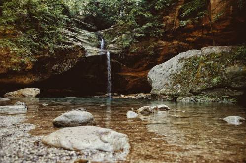 A serene waterfall cascades into a clear pool, surrounded by rocky terrain and lush greenery.