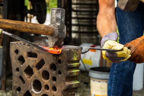A blacksmith hammers a glowing piece of metal on an anvil, sparks flying in a workshop setting.