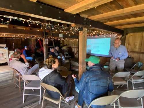 A group of people seated in a barn, listening to a speaker presenting on a screen, with festive lights in the background.