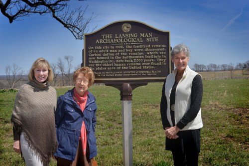Three women stand beside a historical marker for the Lansing Man Archaeological Site on a sunny day.