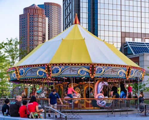 A colorful carousel with a yellow and white canopy, surrounded by people in an urban park setting.