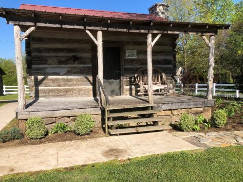 A rustic log cabin with a red roof, wooden steps, and a porch bench, surrounded by greenery and a stone pathway.