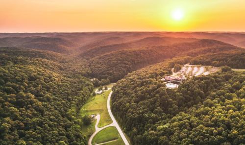Aerial view of rolling green hills at sunset, with a winding road and a building nestled in the valley.