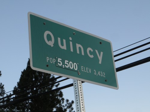 Sign for Quincy, indicating a population of 5,500 and an elevation of 3,432 feet, with trees and power lines in the background.